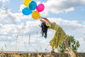 Fototapeta na wymiar A young brunette girl in a green dress is dancing on the ruins against the background of the summer sky with multi-colored balloons in her hands.