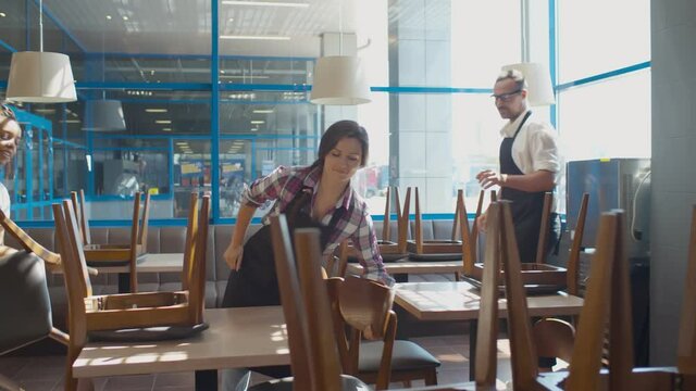 Cafe waiters smile and chat raising chairs to arrange in preparation for closing coffee shop