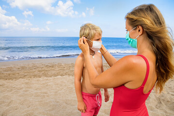 Fototapeta na wymiar Mother put medical mask on child on sea beach. New rules to wear cloth face covering at public places. Cancelled cruise, tour due coronavirus COVID 19. Family vacation, travel lifestyle at summer 2020