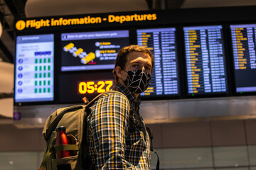 Photo of a guy with a backpack looking at the Departure screen at the airport ready to catch a flight and wearing a reusable face mask.