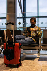 Fototapeta na wymiar Photo of a young and attractive man wearing a shirt in the airport waiting to catch a flight with the suicase ready. Checking his phone and wearing a reusable face mask. Smart casual style