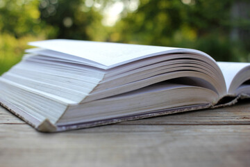 Open old book with flipping book pages on wooden table outdoors. Close-up. Selective focus. Enjoying summer reading. Natural blurred background
