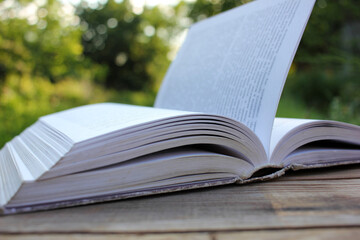 Open old book with flipping book pages on wooden table outdoors. Close-up. Selective focus. Enjoying summer reading. Natural blurred background
