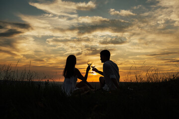 Plakat Silhouette of couple in love at amazing sunset. They drinking white wine and sitting in a meadow. Summer vacation, having fun, positive mood, romantic, silhouette, happy. Copy space.