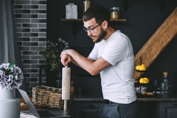Young pensive caucasian bearded man is looking at food recipes in a laptop, while standing in the kitchen, holding a rolling pin in hands. Cooking preparation.
