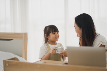 Love of young Asian mother and daughter give a cup of coffee to mom when working by computer laptop in living room at home which smiling and felling happy. Asian love family live at home concept.