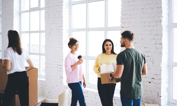 Attractive male and female friends talking while unpacking carton boxes with stuff and goods for new office standing near window in modern loft interior space, employees communicate during relocation