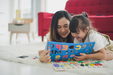 Fototapeta premium Love of young Asian mother and daughter lie on floor and enjoy to play a game near sofa in living room at home which smiling and felling happy. Asian love family live at home concept.