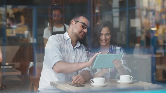 View through window of smiling young couple looking at digital tablet with coffee cups in cafe