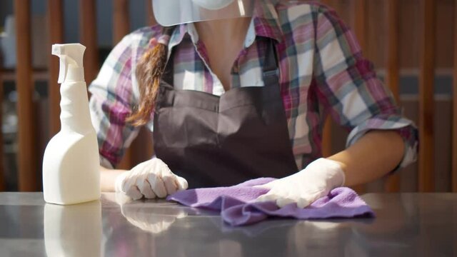 Waitress in n medical and plastic face mask and gloves disinfects surfaces.