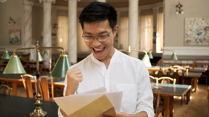 Fototapeta na wymiar Portrait of asian student joyfully opening envelope with exam results in university library. Young attractive guy rejoicing exam results in campus