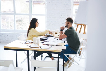 Skilled male and female colleagues dressed in casual wear ordering documentation sitting at table in new office indoor, creative coworkers having brainstorming session collaborating for project