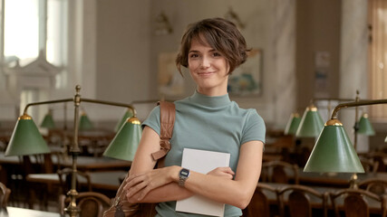 Portrait of beautiful student girl happily looking in camera standing with backpack and textbook in college library