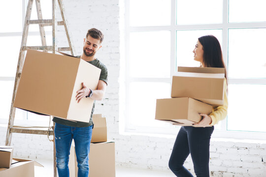 Young smiling couple in love carrying boxes with stuff relocating to apartment talking to each other planning design for room, male and female colleagues packing cardboard boxes moving in new office
