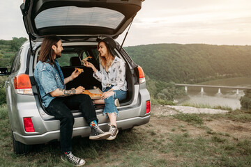 Young Stylish Traveling Hipsters Having Fun Sitting in Car Trunk and Eating Pizza, Travel and Road Trip Concept