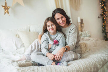 A beautiful woman and a little cute girl, mom and daughter in gray pajamas are sitting on the bed, looking at the camera. Cozy tender photo, Christmas morning concept, scandy style, soft focus.