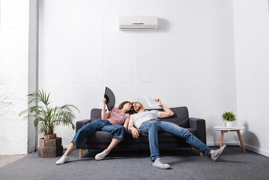 young couple with hand fan and newspaper suffering from heat at home with broken air conditioner
