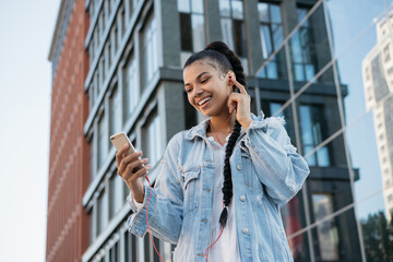 Happy stylish girl holding smartphone, listening to music, smiling outdoors. Beautiful African American woman using mobile phone, communication, looking at digital screen, walking on the street