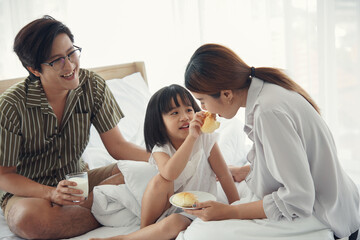 Asian family little daughter give her mom sausage bread on mouth while dad holding a cup of milk...