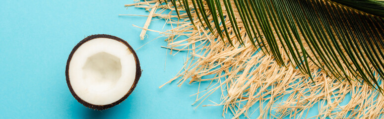 top view of green palm leaf, coconut half, straw hat on blue background, panoramic shot