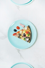 Slice of pizza with vegetables. Dish with pesto sauce, cherry tomatoes, cucumber, Feta cheese, ham, olives and Spanish onion on black dough. ingredients on turquoise serving plate, marble background.
