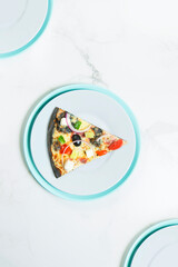Slice of pizza with vegetables: , cucumber, cherry tomatos, olives, ham, Feta cheese, Spanish onion on black dough. Gray and turquoise serving plates, three tablewares on white marble background.
