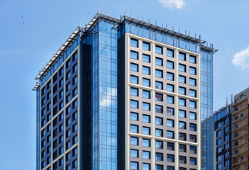 A fragment of the glass facade of a modern concrete building under construction with a reflection of the blue sky and a tower crane.