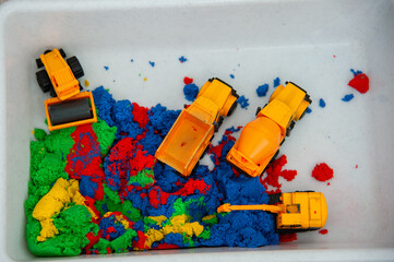 Toy plastic construction machines (skating rink, excavator, dump truck, concrete mixer) in a plastic sandbox with colorful (red, yellow, blue) kinetic sand. View from above.