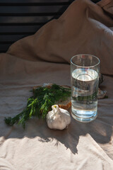 A transparent glass with water, a head of garlic, dill on a slice of bread on a sand-colored linen tablecloth, on a sunny day. Behind a glass are sea shells and gray granite stone. Copy space