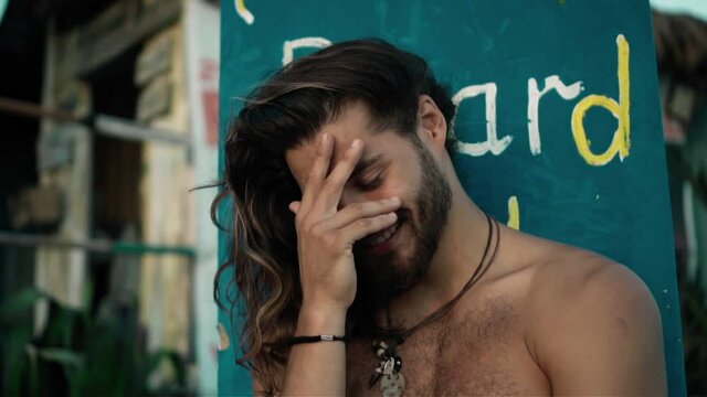 Young handsom European guy brunette with long hair with a wrapped sports body looks forward smiles with an embarrassed look and covers his face with his hand from shame