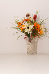 A glass vase with colorful flowers in it. Placed on top of a white wood table next to a white wall. Taken inside with studio bright lights.