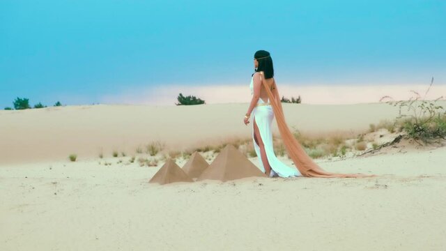 young divine beautiful Egyptian woman goddess Cleopatra walks. White antique vintage dress orange cloak gold jewelry. Girl brunette long loose hair sexy back. blue sky sunny sunset ancient pyramids