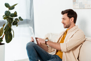 Smiling man sitting on couch and using digital tablet in living room