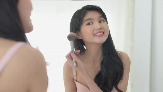 Beauty young asian woman happy and smile with face looking mirror applying makeup with brusher of cheek in the room, beautiful of girl holding brush make-up on facial, skin care and cosmetic concept.