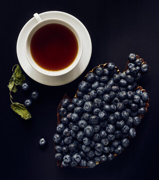 Blueberries on a wooden dish, cup of tea,  and dried melis leaves on a dark background, still life, view from above