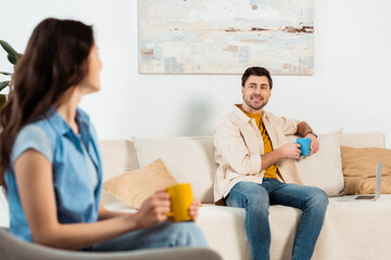 Selective focus of man holding cup of coffee near laptop and smiling at girlfriend in living room