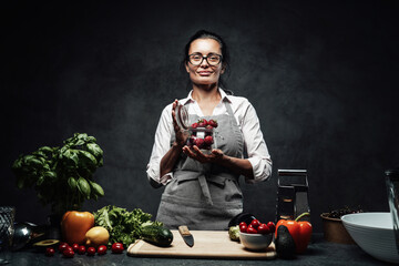 Beautiful middle-aged female chef holds a glass with fresh juicy strawberries next to a table with fresh vegetables in kitchen. Healthy and proper nutrition on a diet. Studio photo on a dark