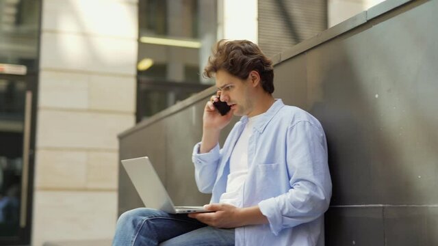 Side view tracking shot of young man using laptop computer and answering phone call outside. Businessman discussing work on cell phone and finishing call sitting on bench outdoors