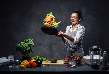 Happy middle-aged female chef tossing chopped vegetables in the air from a frying pan. Healthy food concept. Studio photo on a dark background