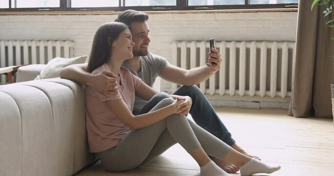 Happy young family couple holding phone looking at screen taking selfie sit on floor in modern room apartment, smiling man and woman embracing making self portrait picture on smartphone at home