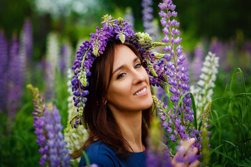 Woman in blue dress in purple lupines field. Meadow of violet flowers in the summer. Girl with long hair holding a lupine bouquet on nature background. Sunset light. Summertime positive scene.