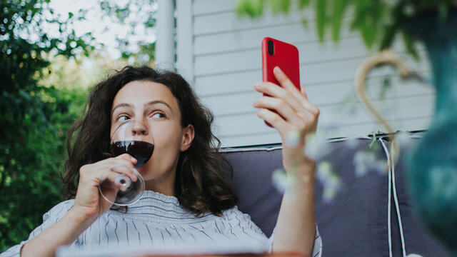 Brunette Woman Drinking Red Wine And Toasting With Best Friend By Video Chat.Girl Making Video Call With Smartphone Lying On Sofa Veranda Outdoor.Video Conference Party Online Meeting.Isolation