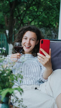 Happy Young Smiling Caucasian Woman Making Video Call With Smartphone Sitting On Sofa Terrace At Home Outdoor. Best Friends Toasting Drinking Red Wine. Video Conference Party Online Meeting. Vertical