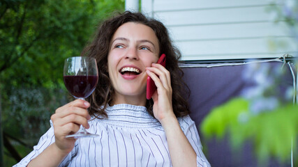 Happy smiling girl enjoying pleasant talk by smartphone with glass of red wine at home terrace sitting on sofa.Young millennial caucasian woman in white summer cloth talking on mobile phone outdoor