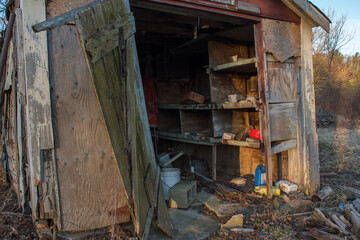 Fototapeta na wymiar Entrance to an abandoned shed with door hanging off and strewn household items and garden chemicals amidst the debris in rural New England.