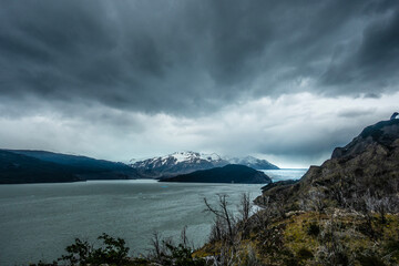 A lake and glacier are seen with snowy mountains and ominous, gloomy clouds. Trees and other foliage as well as rocks appear in the foreground. Torres del Paine National Park, Patagonia, Chile