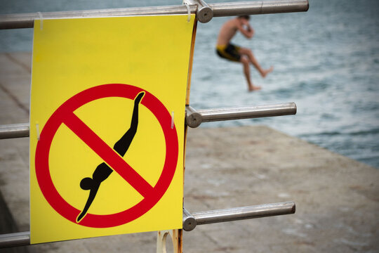 A sign prohibiting diving from the pier against the background of a blurry figure of a young man jumping the pier.