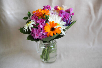 in a transparent glass vase is a bouquet of bright summer flowers in white, pink, orange on a beige textured background side view