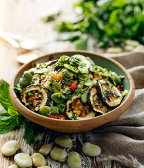 Broad bean salad with the addition of various vegetables sprinkled with fresh herbs in a ceramic bowl on a  wooden table close up view