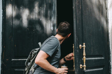 A young caucasian man with backpack opens the big wooden door to the house and looks inside.